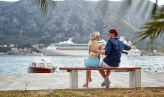 A smiling couple on a bench with a cruise ship in the background, which may have them wondering, "How safe is cruising?".