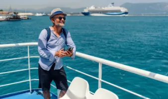Smiling man searching how to get the best deal on a cruise on his phone with a ship in background.