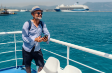 Smiling man searching how to get the best deal on a cruise on his phone with a ship in background.