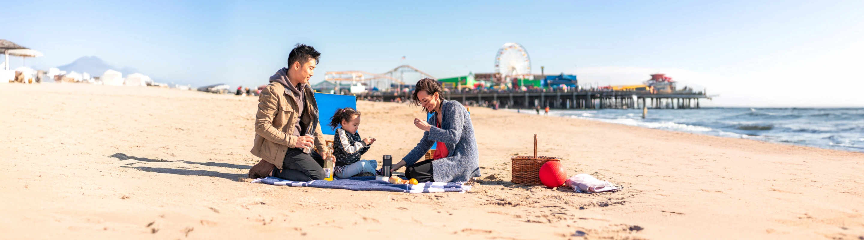 two parents, with travel insurance for California, and their toddler enjoy a picnic on a beach near Santa Monica Pier