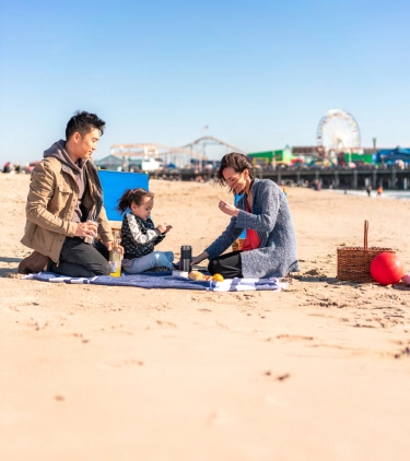 two parents, with travel insurance for California, and their toddler enjoy a picnic on a beach near Santa Monica Pier