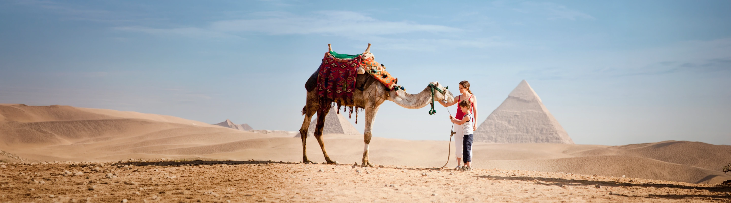 a mother-son duo with travel insurance for Egypt pet a camel in a desert with a pyramid in the background