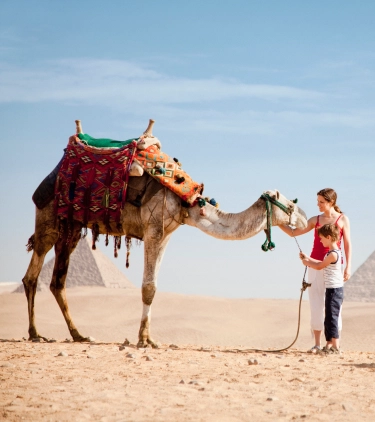 a mother-son duo with travel insurance for Egypt pet a camel in a desert with a pyramid in the background