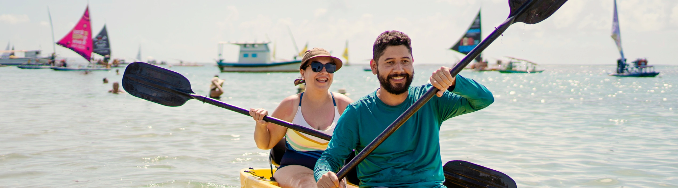 a young couple with adventure travel insurance tandem kayak in the ocean with sailboats in the background