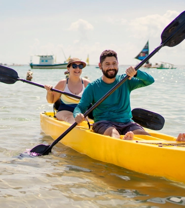 a young couple with adventure travel insurance tandem kayak in the ocean with sailboats in the background