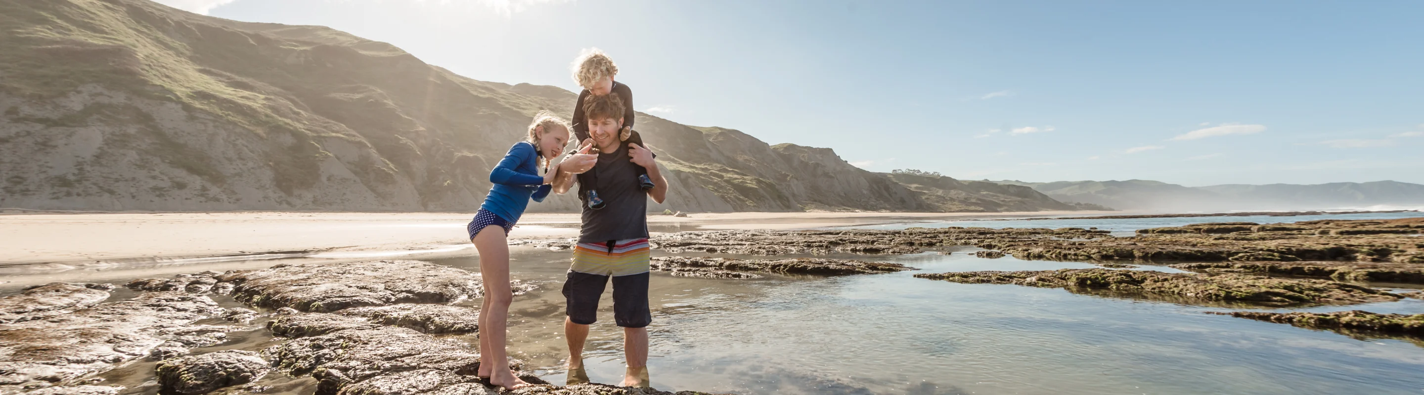 a father with his two kids (one on his shoulder), on the East Cape coastline with travel insurance for New Zealand