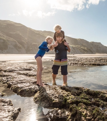 a father with his two kids (one on his shoulder), on the East Cape coastline with travel insurance for New Zealand