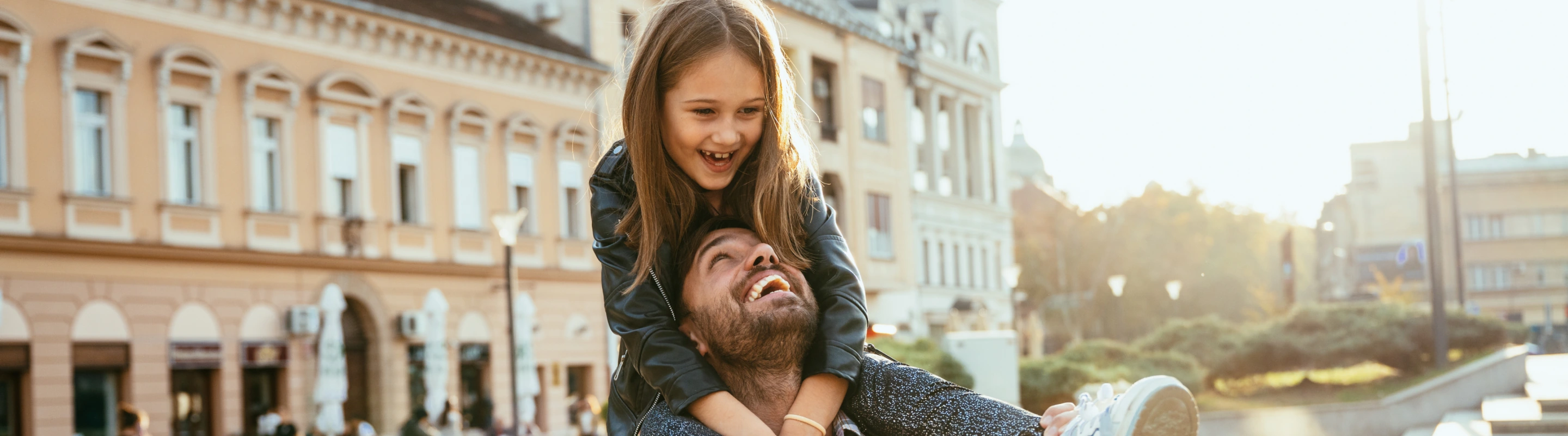 a father with travel insurance for city trips laughs as he holds his daughter on his shoulders nearby classical buildings 