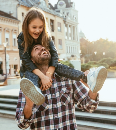 a father with travel insurance for city trips laughs as he holds his daughter on his shoulders nearby classical buildings 