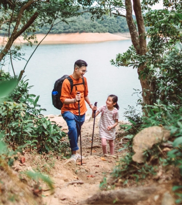 Father with comprehensive travel insurance smiling at his daughter while hiking in the woods with a lake in the background