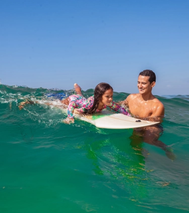 Young girl on surfboard in the ocean while her father holds the board with travel insurance for South America