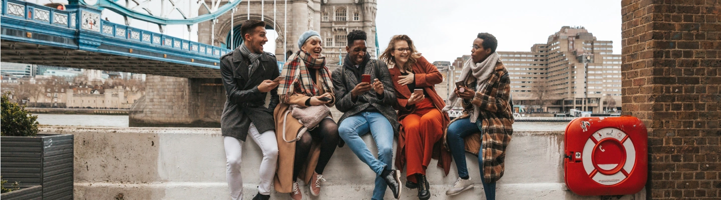 a group of five friends with international travel insurance sit on a ledge, laughing mid-conversation near the London Bridge
