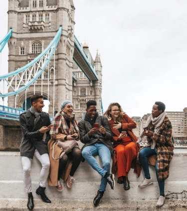 a group of five friends with international travel insurance sit on a ledge, laughing mid-conversation near the London Bridge