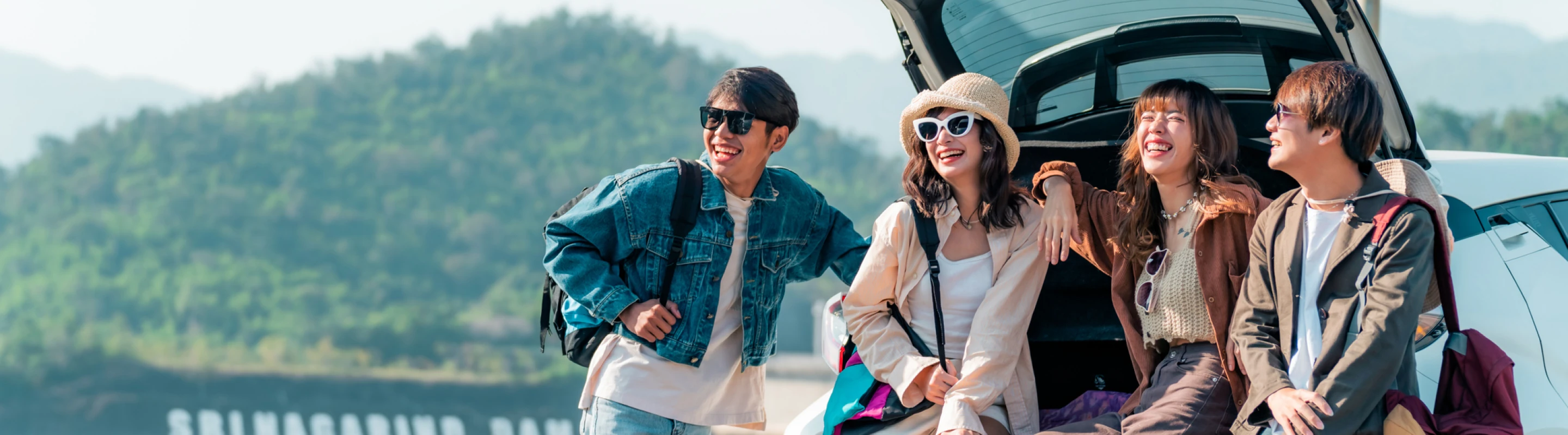 Four young adult friends with group travel insurance sit on the edge of their car’s open trunk during a road‑trip getaway