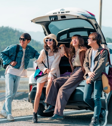 Four young adult friends with group travel insurance sit on the edge of their car’s open trunk during a road‑trip getaway