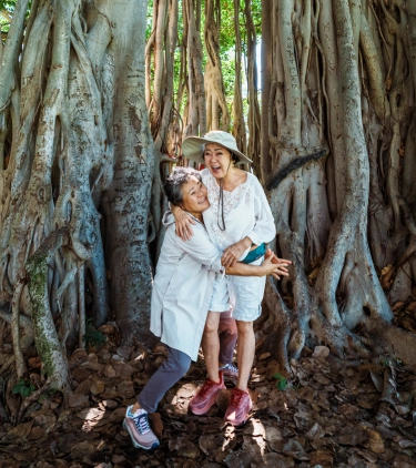 Two female senior friends with travel insurance for Hawaii, hugging and laughing at the base of a banyan tree