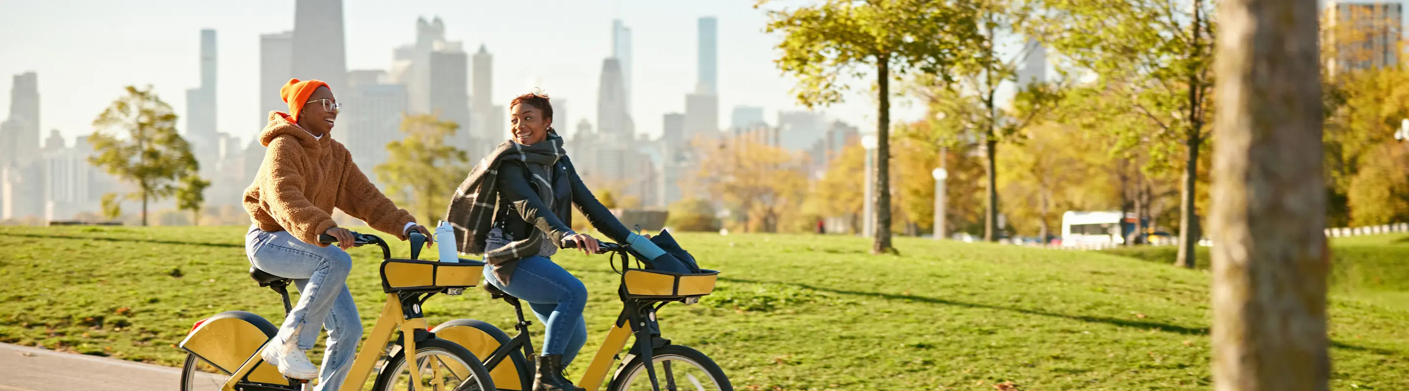 Two female friends with travel insurance for Illinois riding bikes in a park with the Chicago skyline in the background