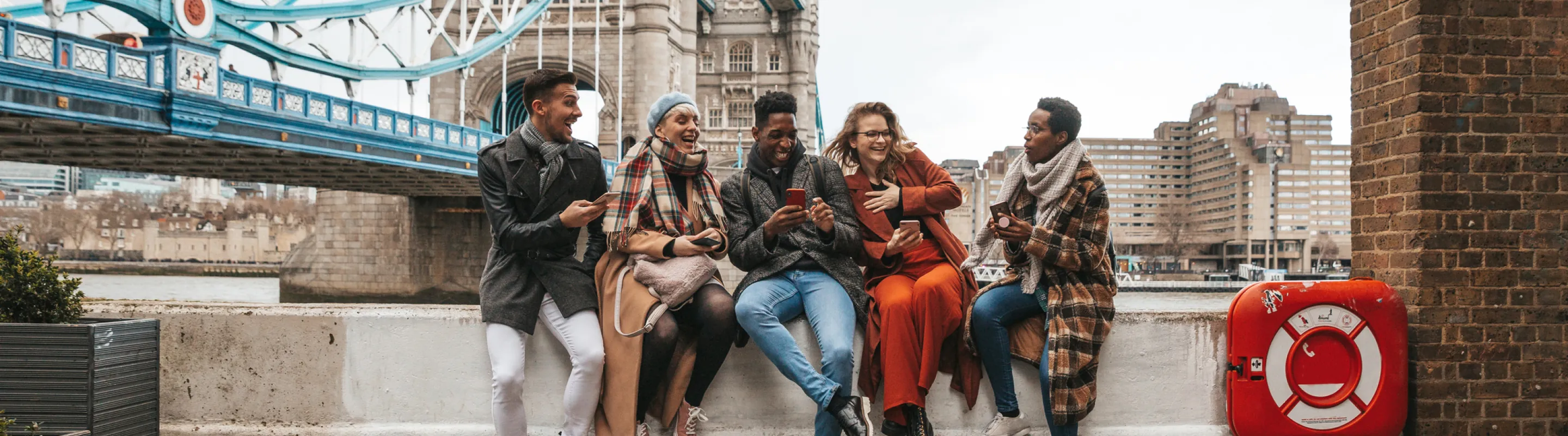 a group of friends with travel insurance for the United Kingdom, laughing as they sit on a ledge near the London Bridge