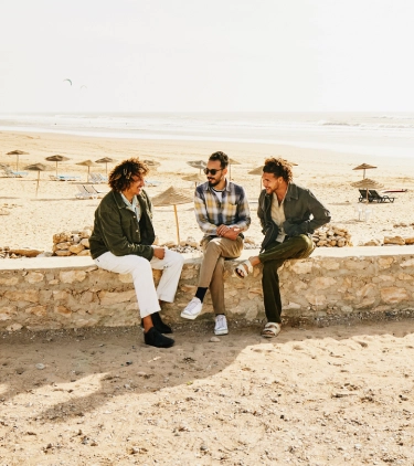 Three adults with last-minute travel insurance sitting on a stone wall and talking with the beach in the background