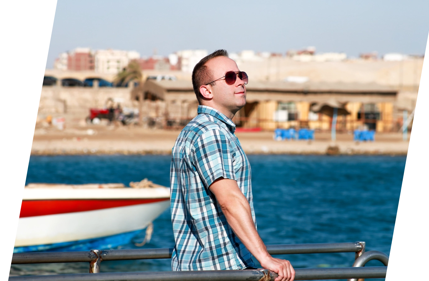 a man with sunglasses, a plaid shirt, and travel insurance for Egypt stands on the water's edge as a boat passes by