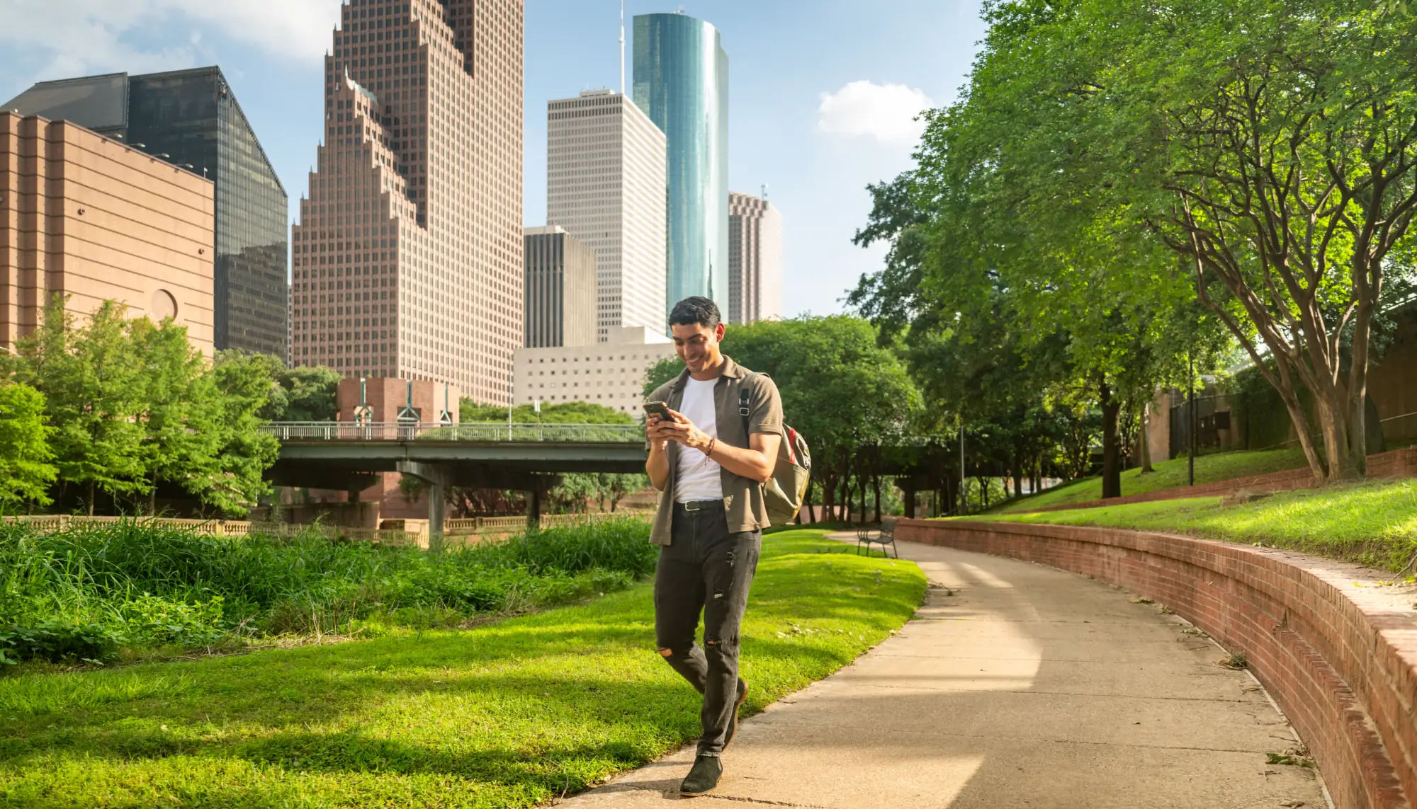 a young man looks at a travel app on his phone as he strolls through a city park in Houston, Texas, on vacation
