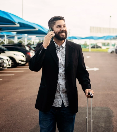 a young man with road trip travel insurance is on his cellphone and rolls his suitcase in an outdoor parking lot 