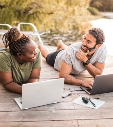 a young man and woman lay on their stomachs on a dock while they work remotely, insured with business travel insurance 