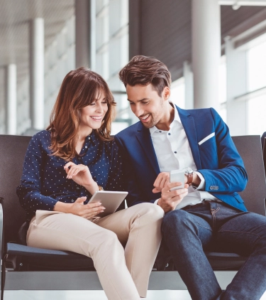 two young professionals look at their work devices as they sit in an airport terminal, insured with business travel insurance 