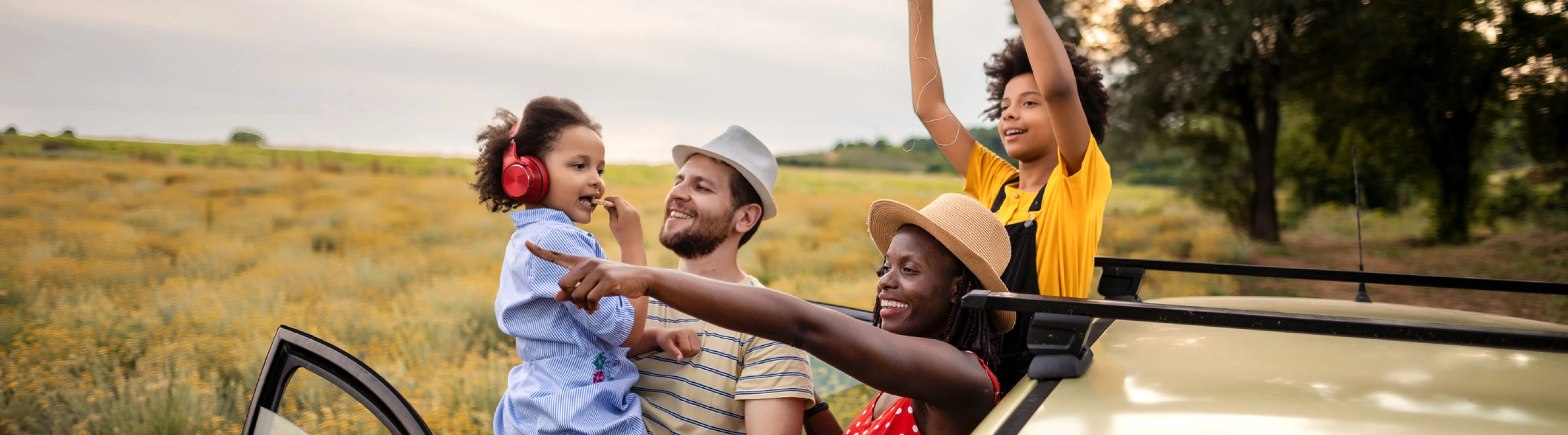 a mixed-race family of four with road trip travel insurance exits their SUV on a dirt road to soak up views of a grassy field 