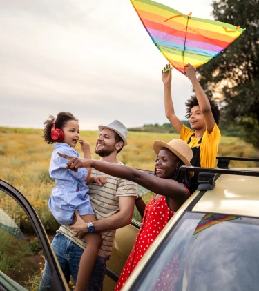 a mixed-race family of four with road trip travel insurance exits their SUV on a dirt road to soak up views of a grassy field 