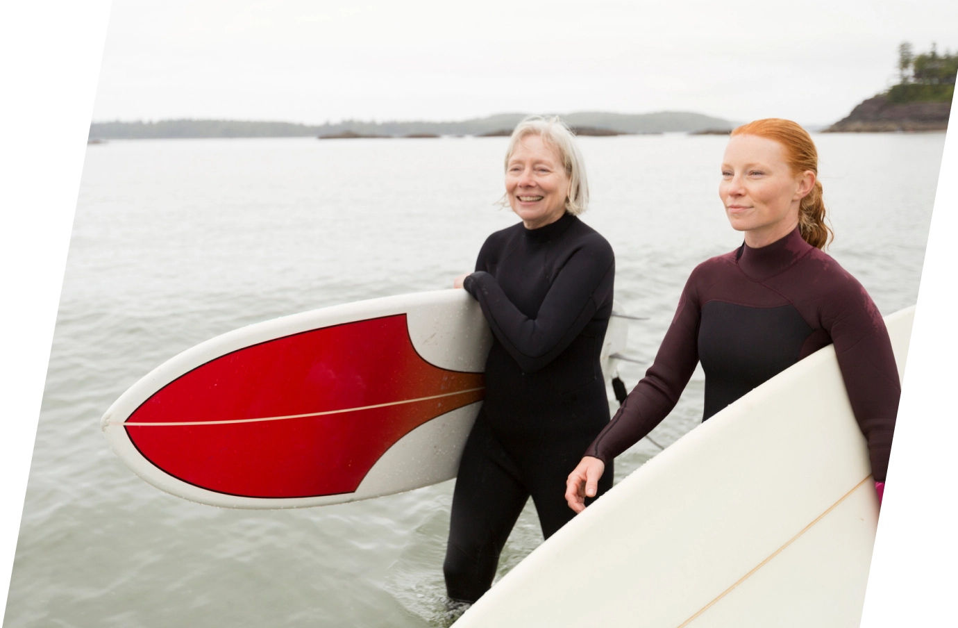 Senior mother and adult daughter with travel insurance for Canada carrying surfboards along the water