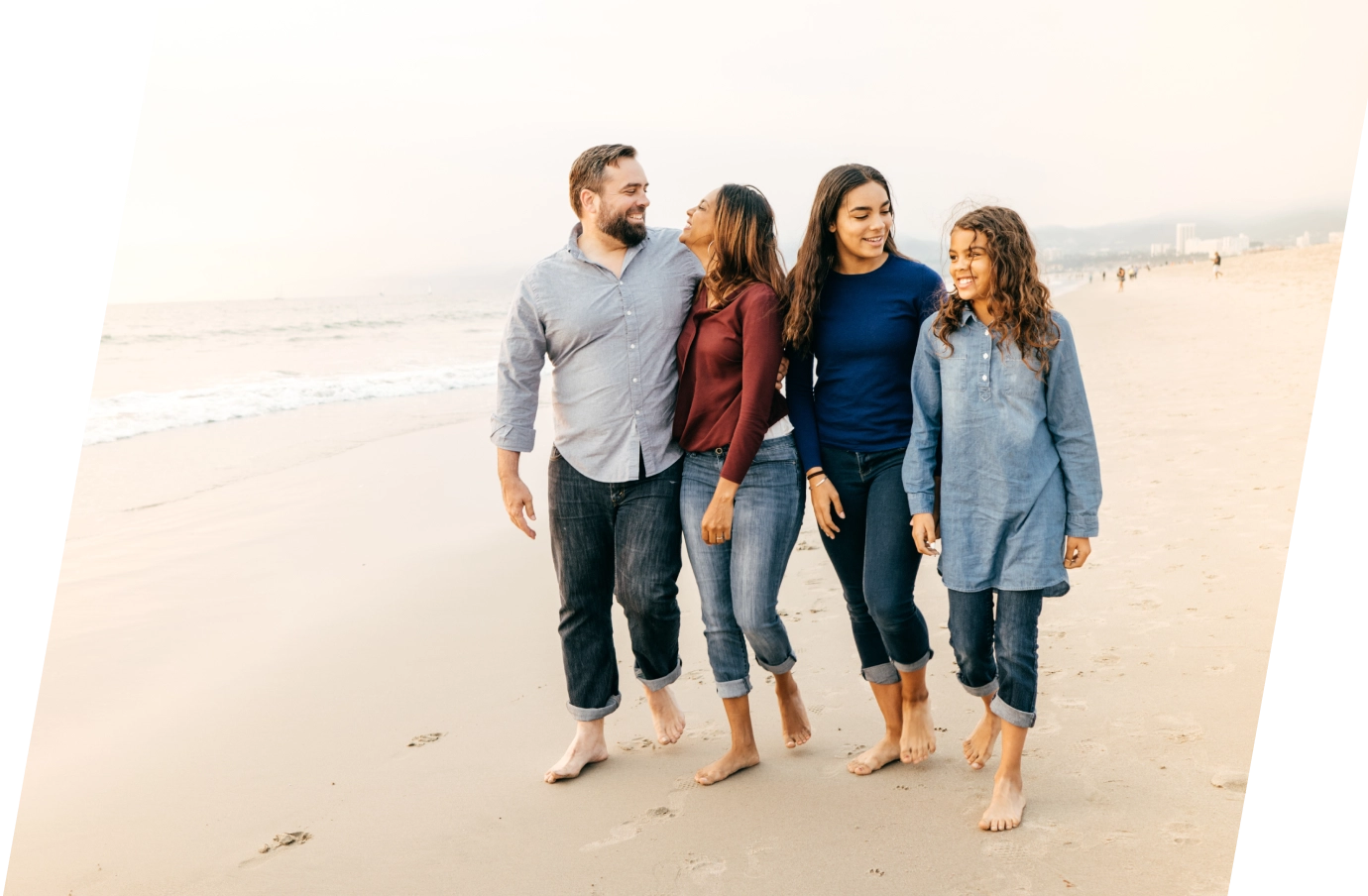 a multiracial family of four takes a walk along a beach barefoot together, insured with travel insurance for California 