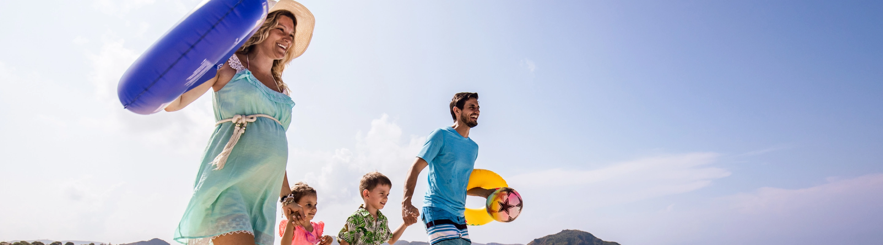 Expecting parents with travel insurance for pregnancy run down a beach while holding hands with their children and carrying inflatable tubes