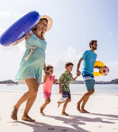 Expecting parents with travel insurance for pregnancy run down a beach while holding hands with their children and carrying inflatable tubes