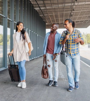 three young adults with group travel insurance walk past an airport’s exterior with their luggage