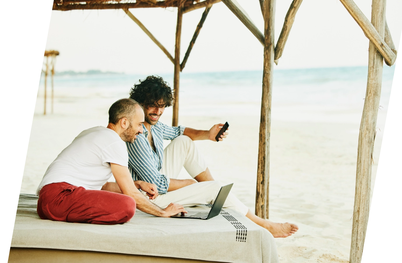 two young men with business travel insurance look at a laptop while sitting in a beach cabana by the ocean 