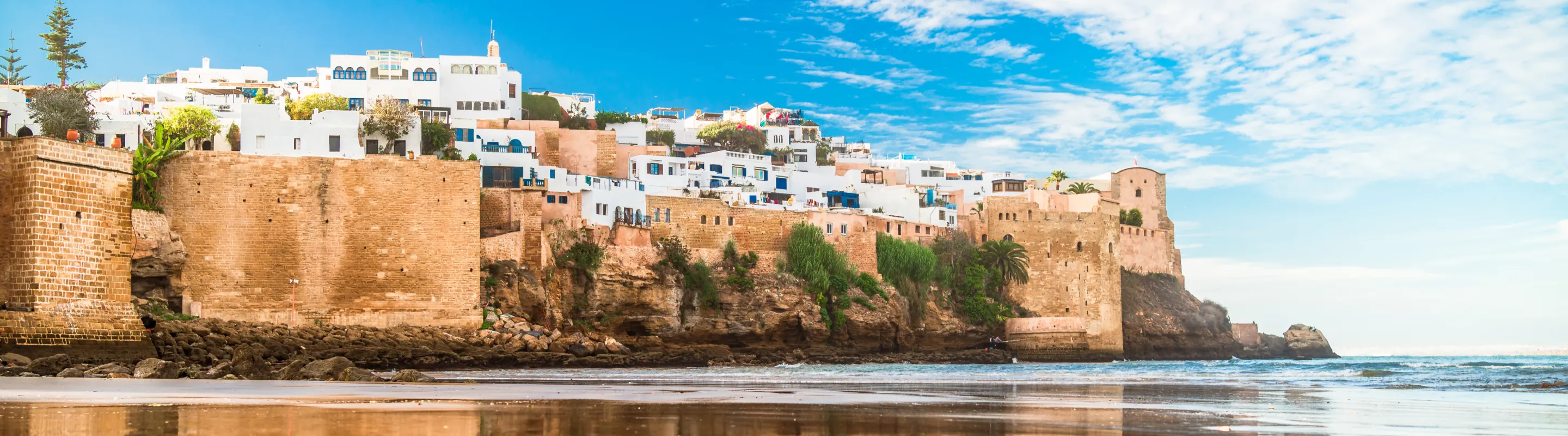 a view of a stone sea wall, a beach, and white-washed buildings in Asilah, where many visit with travel insurance for Morocco