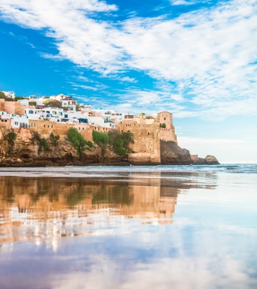 a view of a stone sea wall, a beach, and white-washed buildings in Asilah, where many visit with travel insurance for Morocco