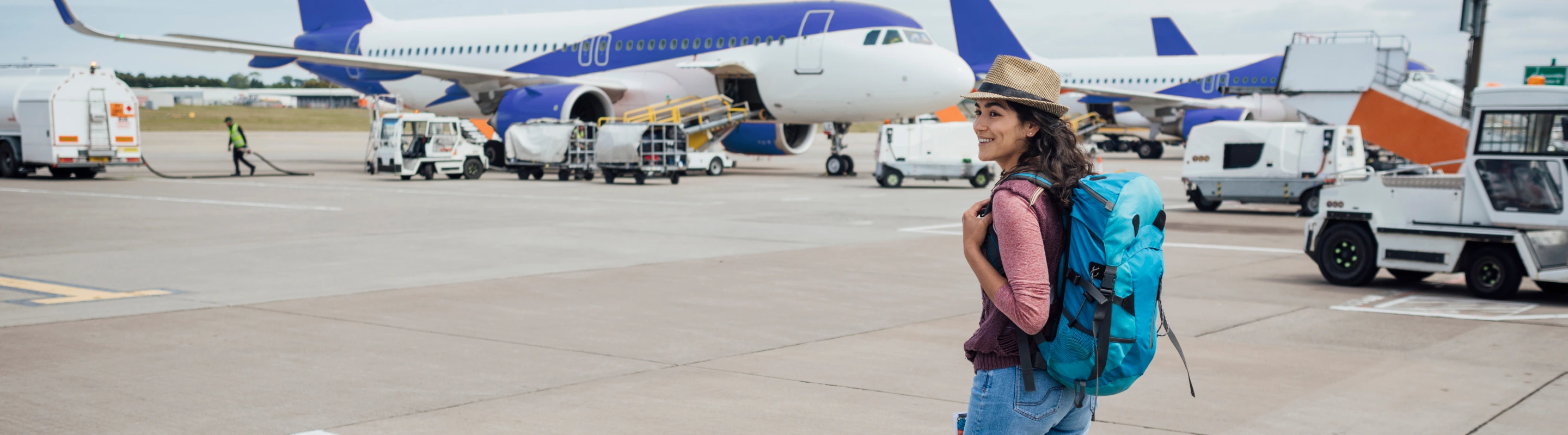 a young woman with a hat and backpacker travel insurance walks across the airport tarmac alongside airplanes