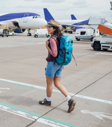 a young woman with a hat and backpacker travel insurance walks across the airport tarmac alongside airplanes