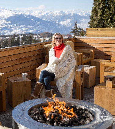 a woman with travel insurance for Switzerland is wrapped in a blanket in front of a fire on a Swiss chalet's balcony