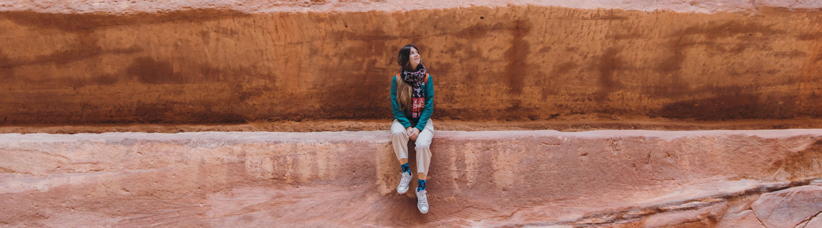 a young woman with a scarf, a turquoise shirt, and travel insurance for Jordan poses on a carved rock ledge