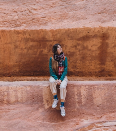 a young woman with a scarf, a turquoise shirt, and travel insurance for Jordan poses on a carved rock ledge