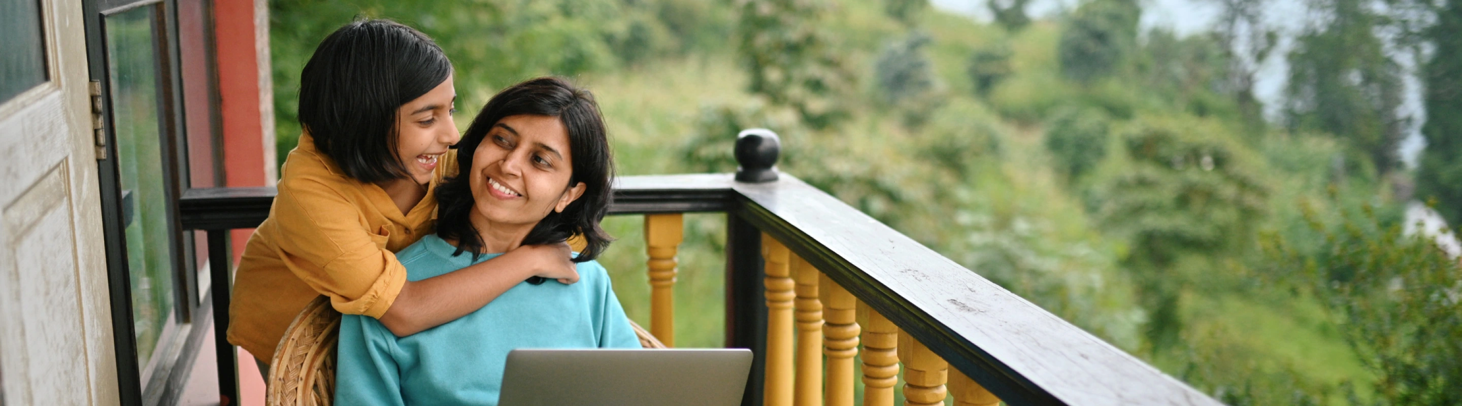 a mother with business travel insurance works on her laptop on a balcony overlooking greenery while her daughter hugs her 
