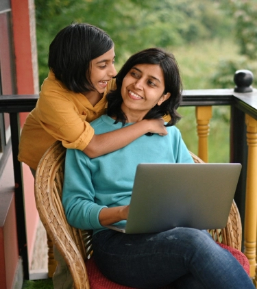 a mother with business travel insurance works on her laptop on a balcony overlooking greenery while her daughter hugs her 