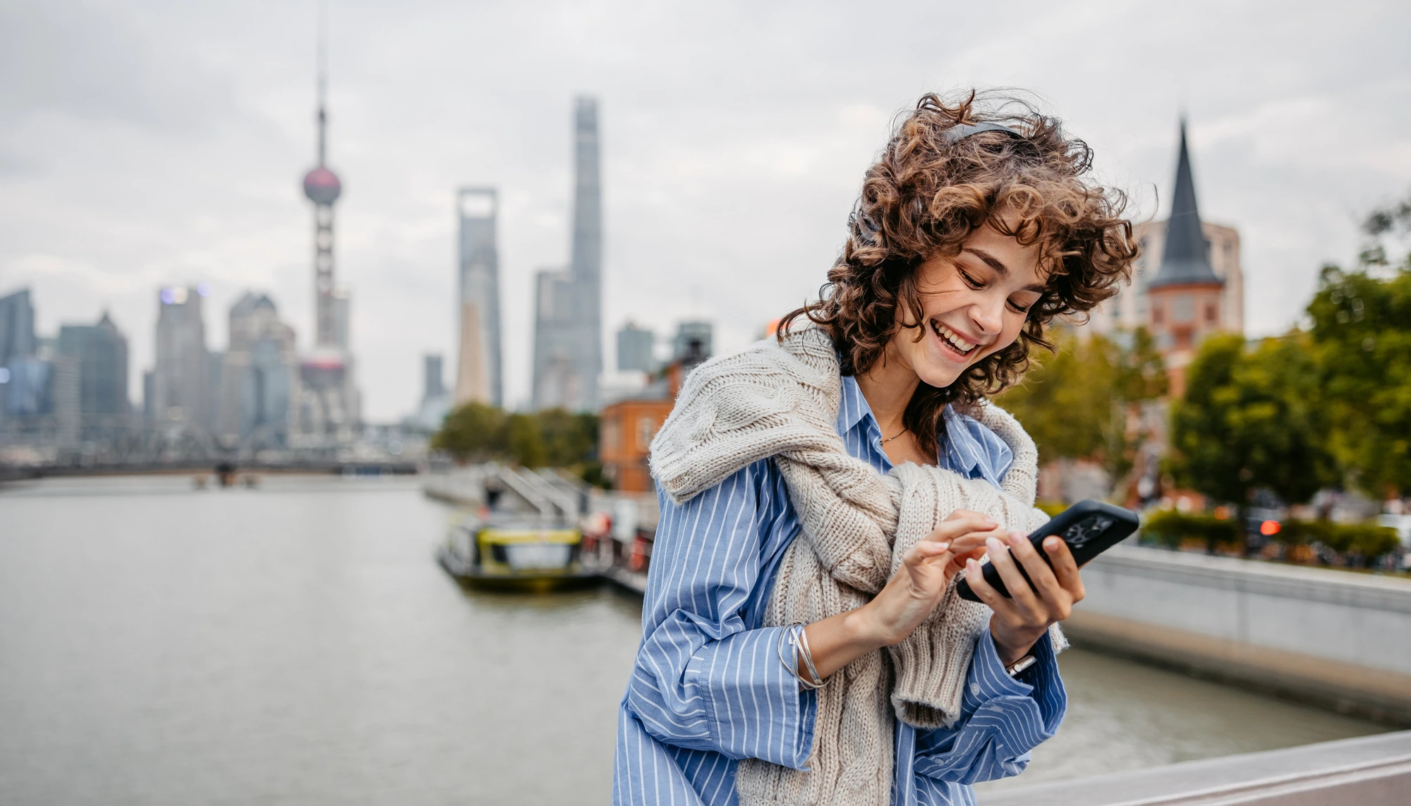 Young woman checking a travel app on her phone while standing on a bridge with Shanghai in the background