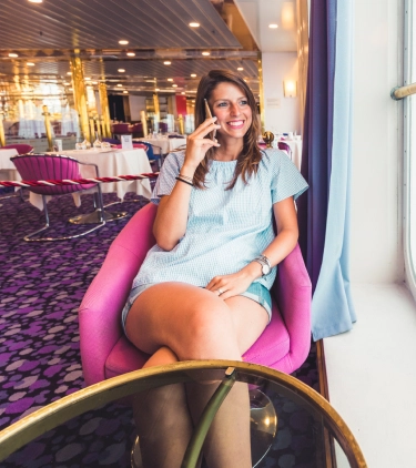 a young woman with cruise travel insurance sits in a hot pink chair by the window in the onboard dining room on her phone