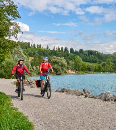 Two middle-aged women with travel insurance for Germany biking along Lake Alpsee in Bavaria