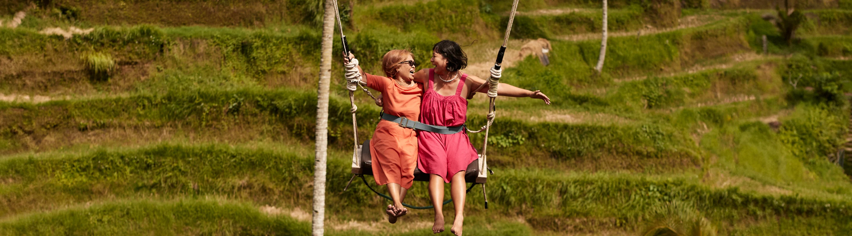 Two women with travel insurance for Bali smiling on a swing in front of rice terraces