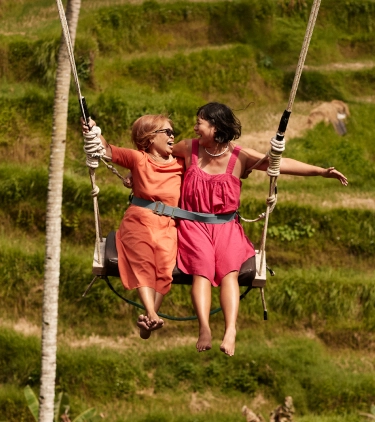 Two women with travel insurance for Bali smiling on a swing in front of rice terraces